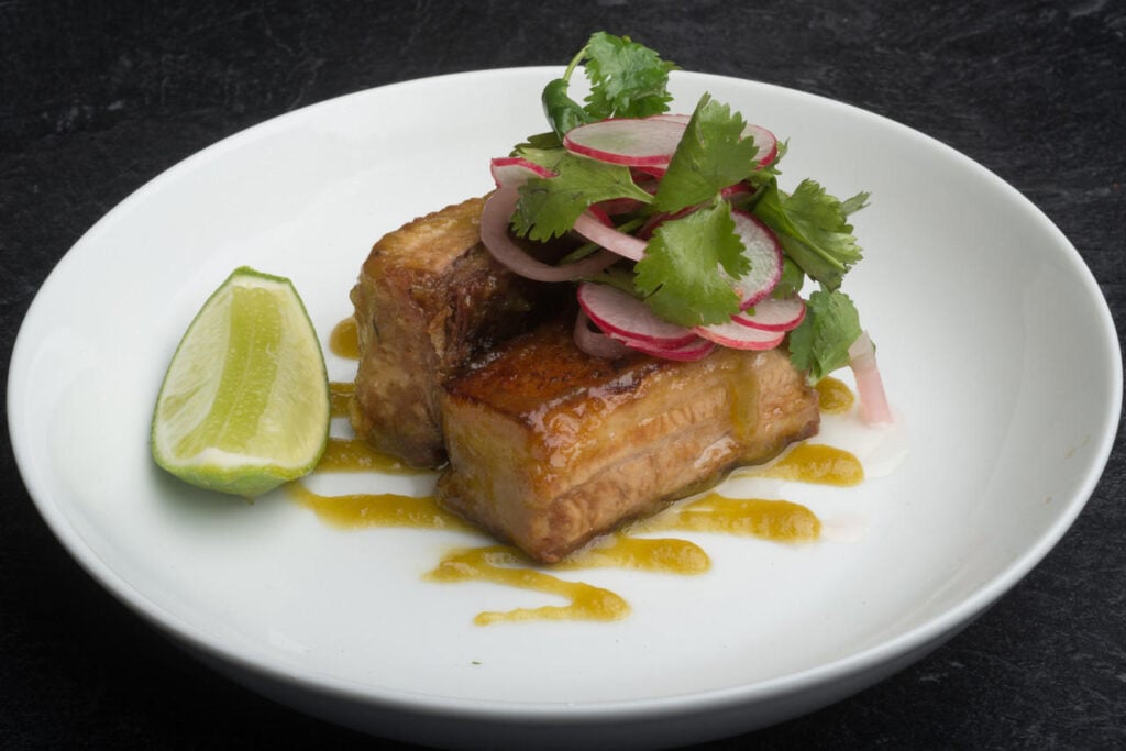 Succulent roasted pork belly topped with fresh cilantro and sliced radishes sits on a white plate, accented by a wedge of lime and yellow sauce, set against a dark textured background in Conejo Valley.