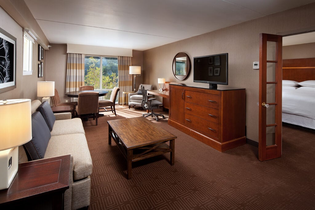 Hotel suite features a cozy seating area with a sofa, armchairs, and a coffee table. A wooden dresser supports a TV.