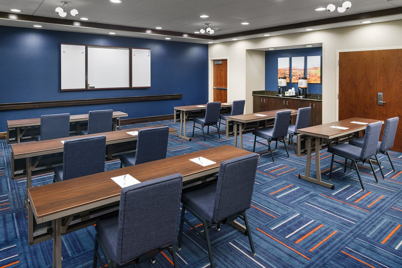 Conference room featuring neatly arranged tables and blue chairs in rows facing a blank whiteboard.