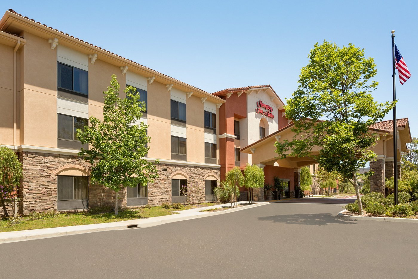 Hampton Inn & Suites building with a tan facade and stone accents, surrounded by trees and greenery.