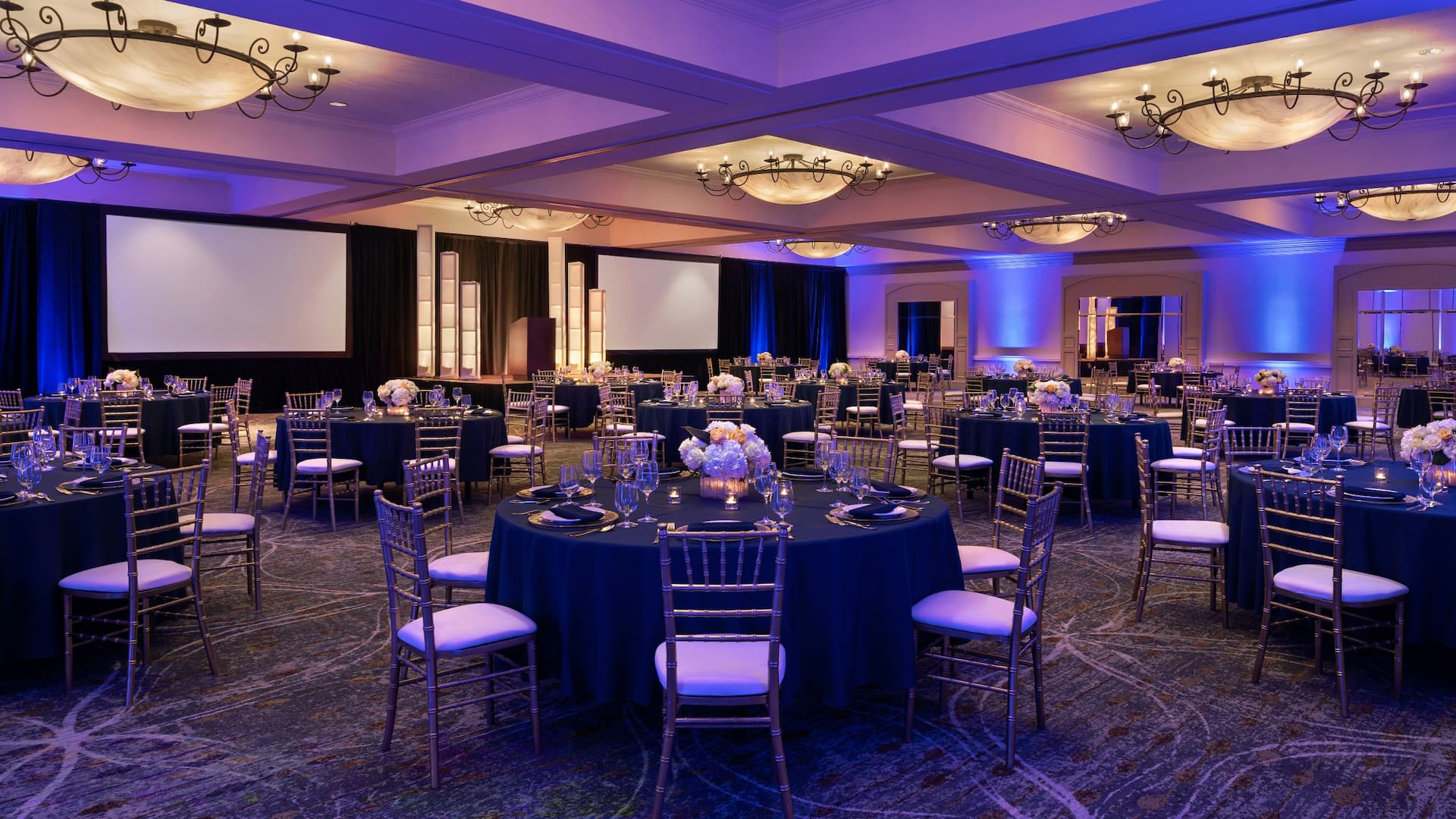 Elegant banquet hall setup features round tables with navy tablecloths, golden chairs, floral centerpieces, and candles.
