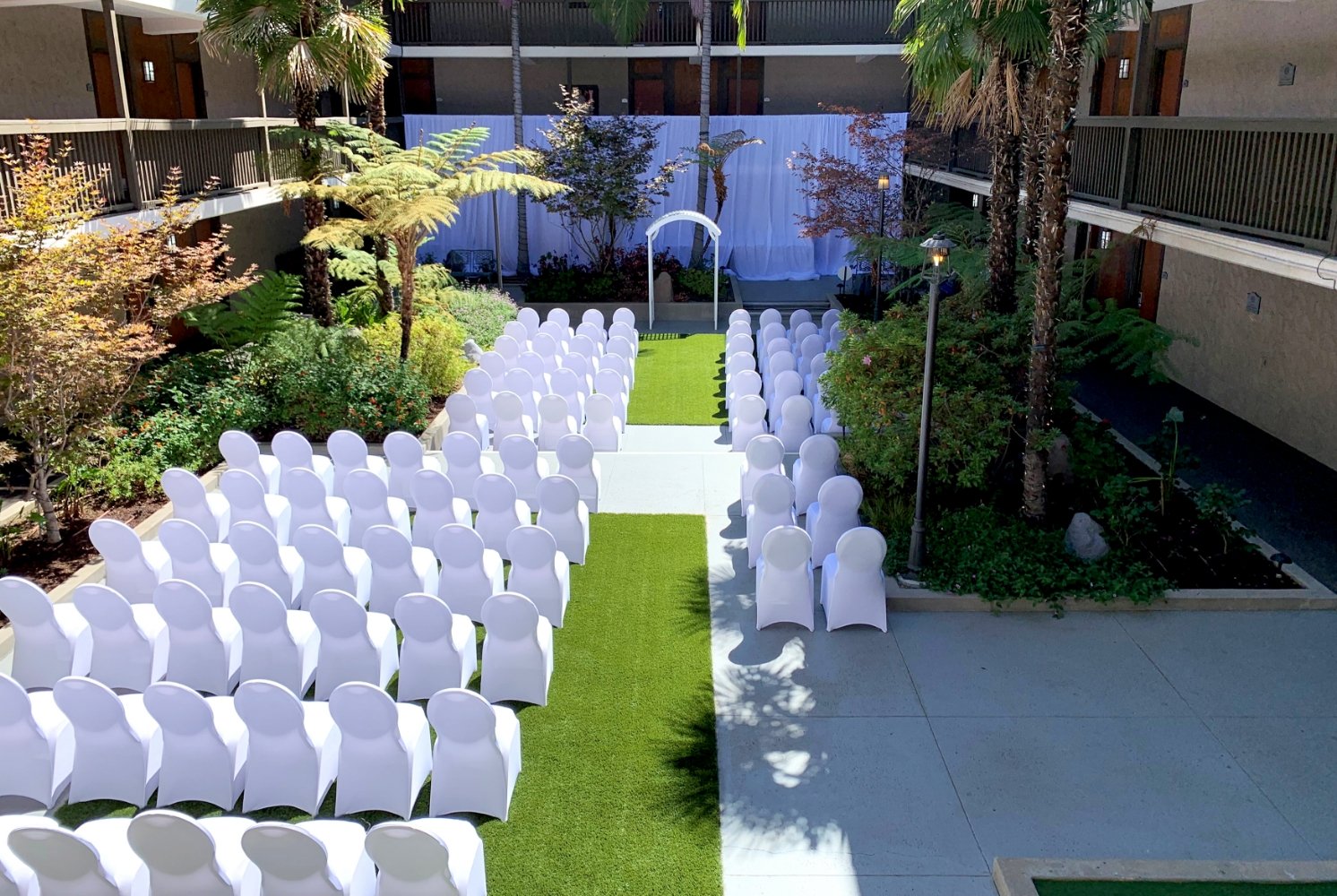 Rows of white covered chairs face a white archway on green artificial grass, surrounded by lush plants and trees in an outdoor courtyard, set for a wedding ceremony in Conejo Valley.