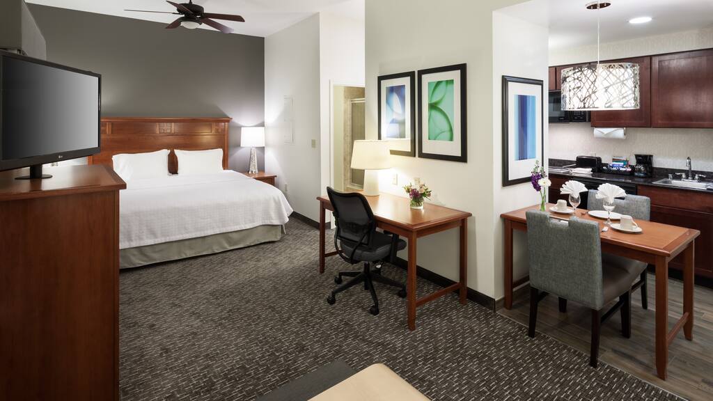 Hotel suite features a king-sized bed with white linens, a flat-screen TV, a work desk, a dining table, and a kitchenette with wooden cabinetry. Modern decor includes framed abstract art in Conejo Valley.