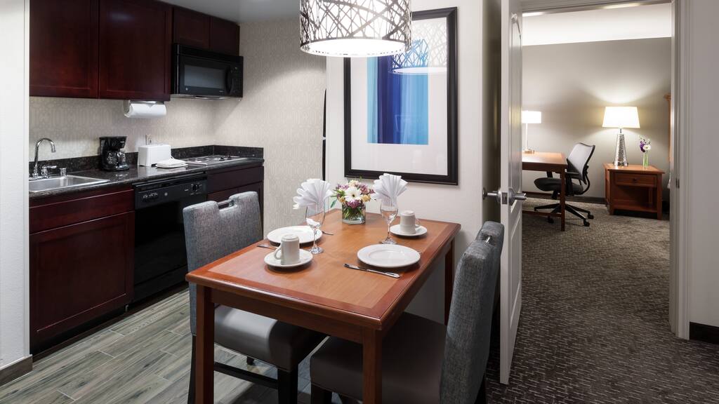 A wooden dining table with chairs set for two is in a modern kitchen, featuring dark cabinets and appliances.