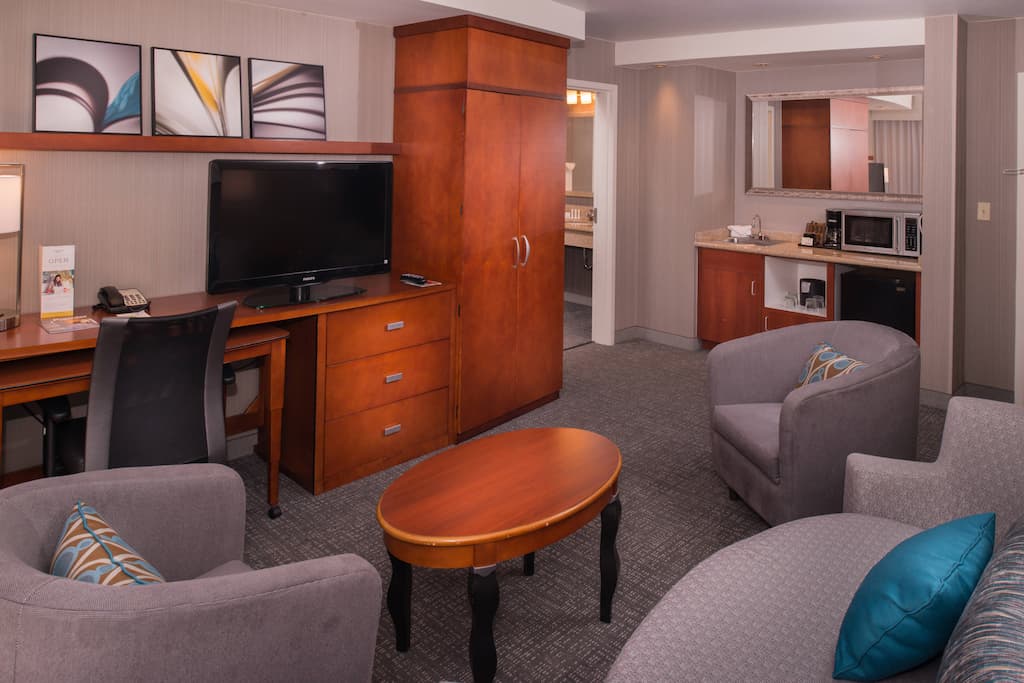 Cozy hotel suite features a flat-screen TV on a wooden desk, armchairs with patterned pillows, and a kitchenette with a microwave and fridge with a comfortable and modern living space in Conejo Valley.