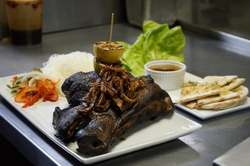 Grilled meat garnished with caramelized onions and skewered lemons sits on a square plate, accompanied by fresh lettuce, a side of pickled vegetables, rice noodles, sauce in a ramekin, and neatly arranged pita bread, set on a stainless steel countertop in Conejo Valley.