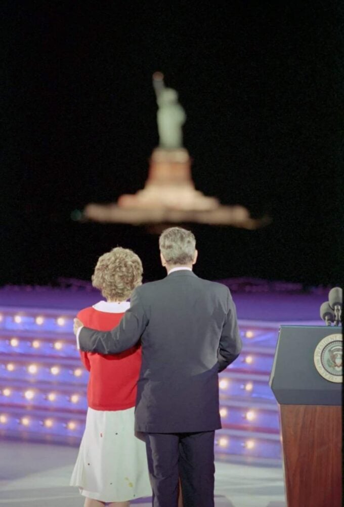 A couple stands facing the Statue of Liberty, illuminated at night. They are on a stage with a podium displaying a seal.