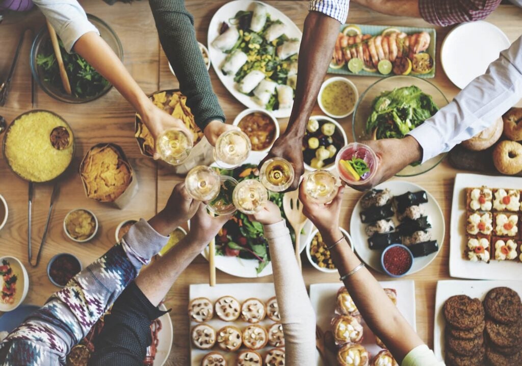 People clinking glasses in a toast over a table filled with diverse dishes including sushi, salads, chips, and desserts with a festive dining atmosphere in Conejo Valley.