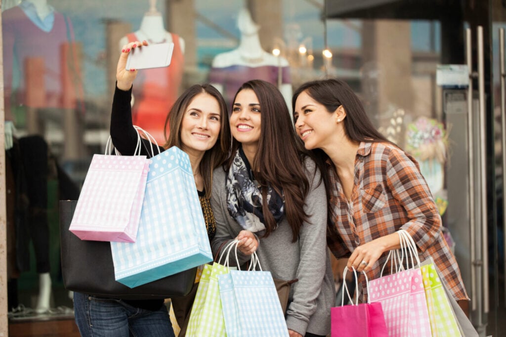 Three women smiling and taking a selfie outside a clothing store.