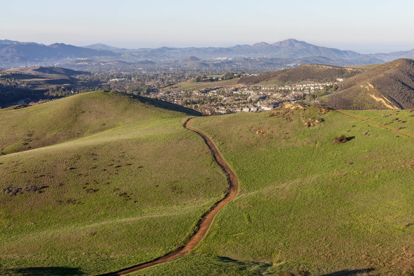 A dirt trail winds through rolling green hills with a suburban neighborhood and distant mountains in the background, capturing a scenic landscape view suitable for hiking adventures in Conejo Valley.