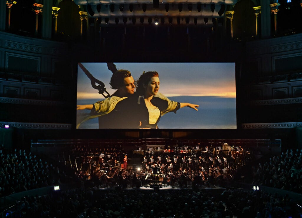 A large screen displays a famous ship scene while an orchestra performs in an ornate concert hall, surrounded by an audience enjoying the movie-concert experience in Conejo Valley.