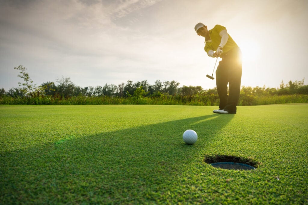 Golfer performs a putt on a sunny green field, aiming at a nearby hole, surrounded by trees under a cloudy sky.