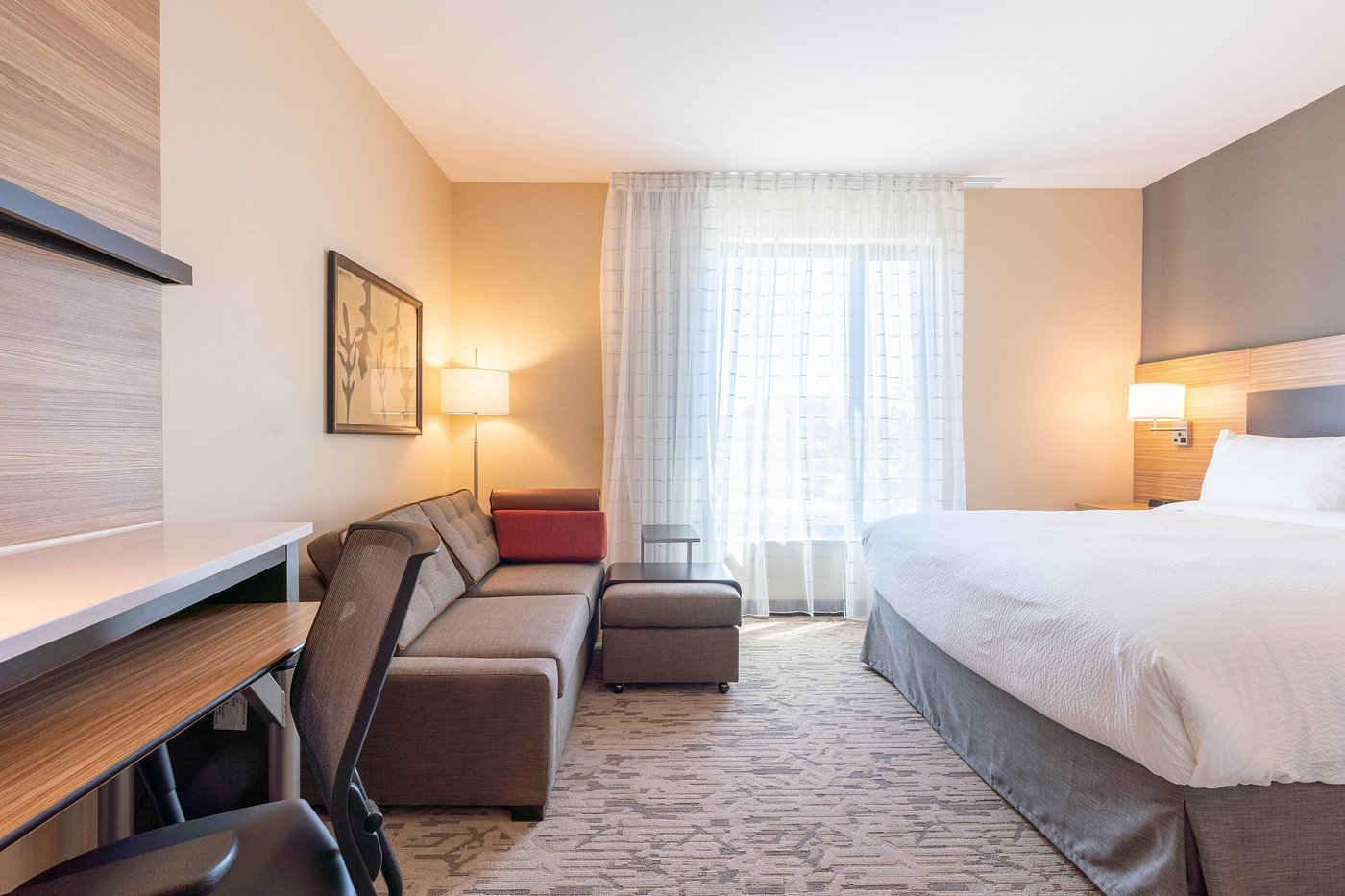 Hotel room features a cozy bed with white linens beside a gray sofa adorned with red cushions.