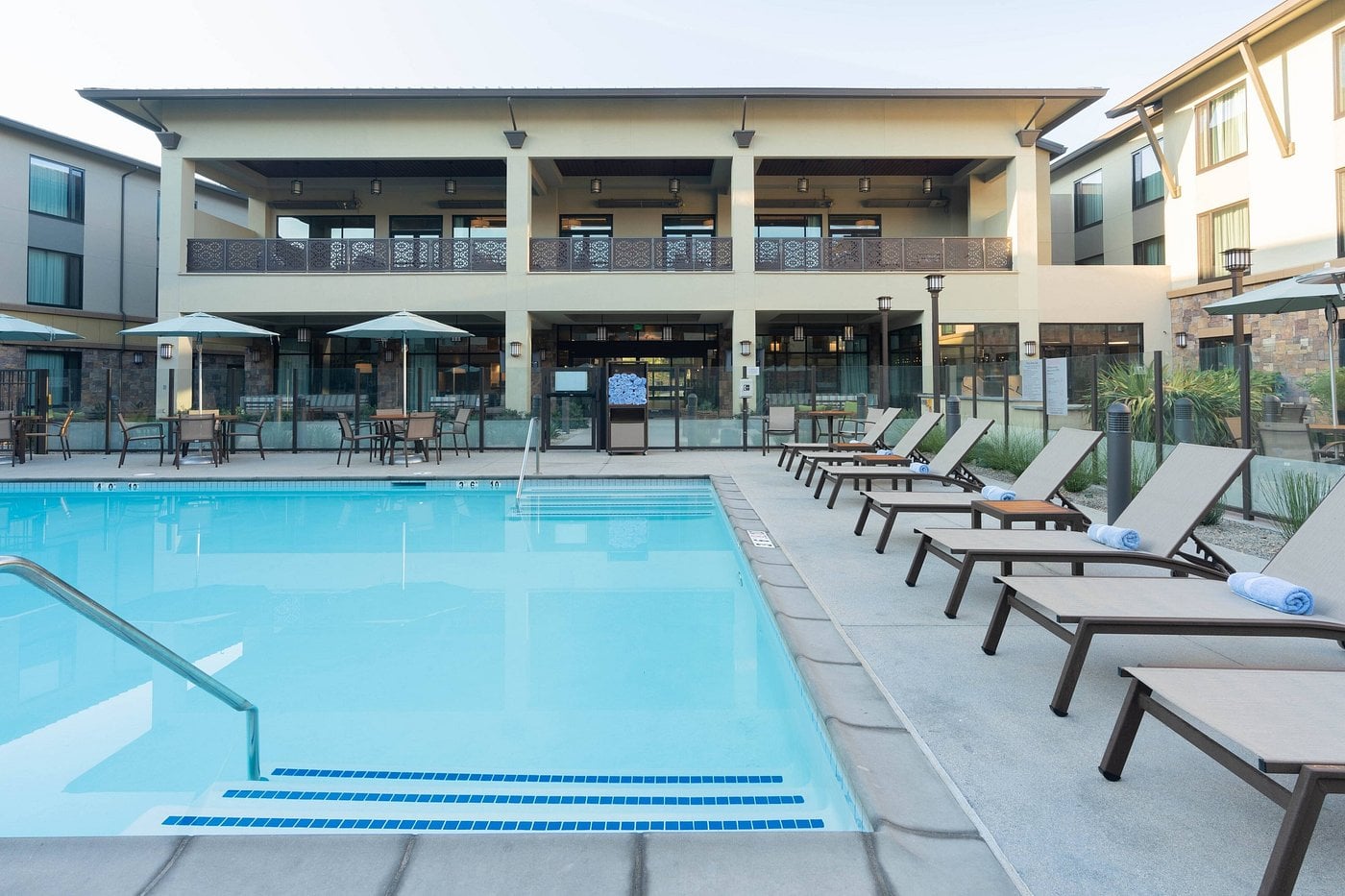Outdoor swimming pool surrounded by lounge chairs and umbrellas; situated in a modern, upscale hotel courtyard with two-story balconies and large windows, ideal for relaxing in a resort setting in Conejo Valley.