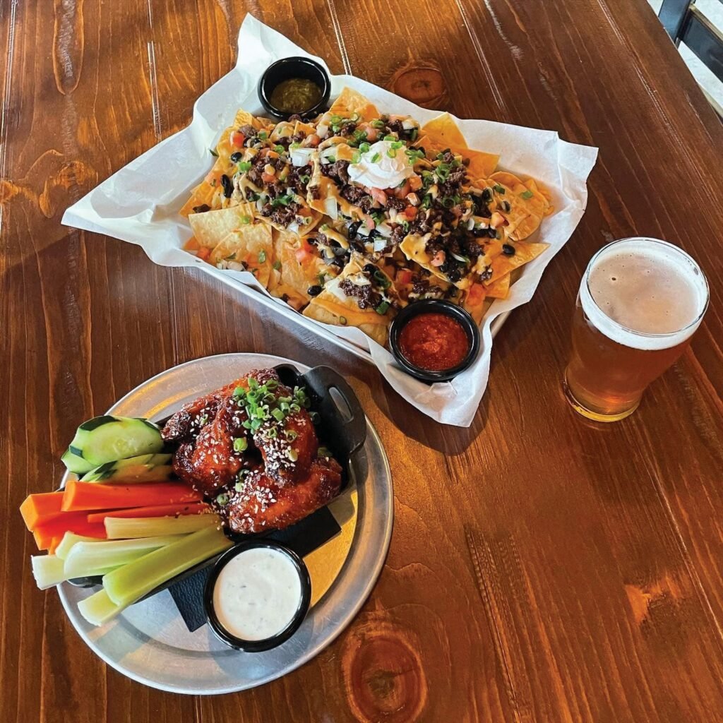 A wooden table features loaded nachos with toppings and dipping sauces, next to a plate of saucy chicken wings, vegetables, and ranch.