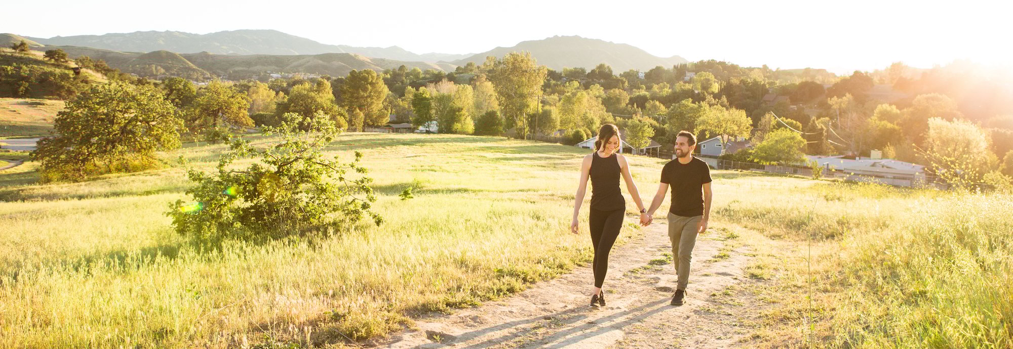 A couple walks hand-in-hand along a sunlit path in a grassy field, surrounded by trees and distant hills under a bright sky in Conejo Valley.