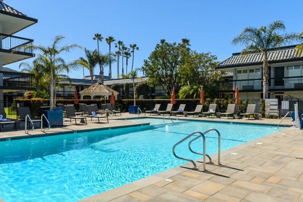 Outdoor swimming pool with clear blue water and surrounding lounge chairs, umbrellas, and palm trees.