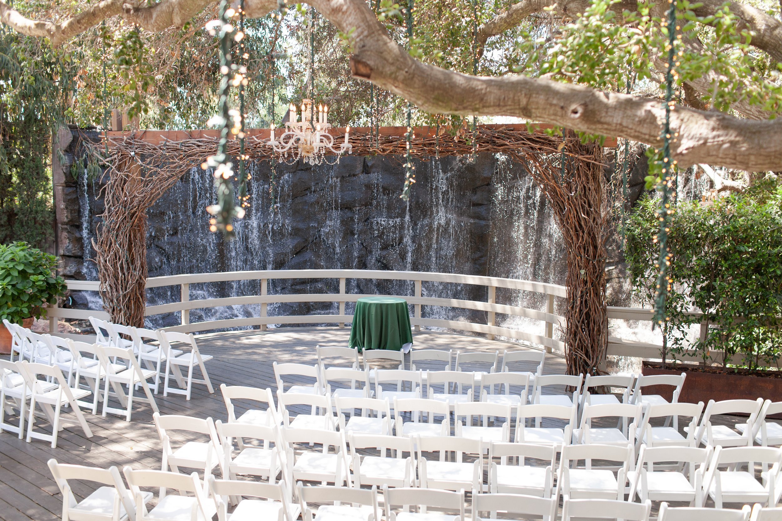 White chairs arranged facing a rustic wooden arch adorned with a chandelier, set in front of a cascading waterfall surrounded by lush greenery. Ideal outdoor wedding venue in Conejo Valley.