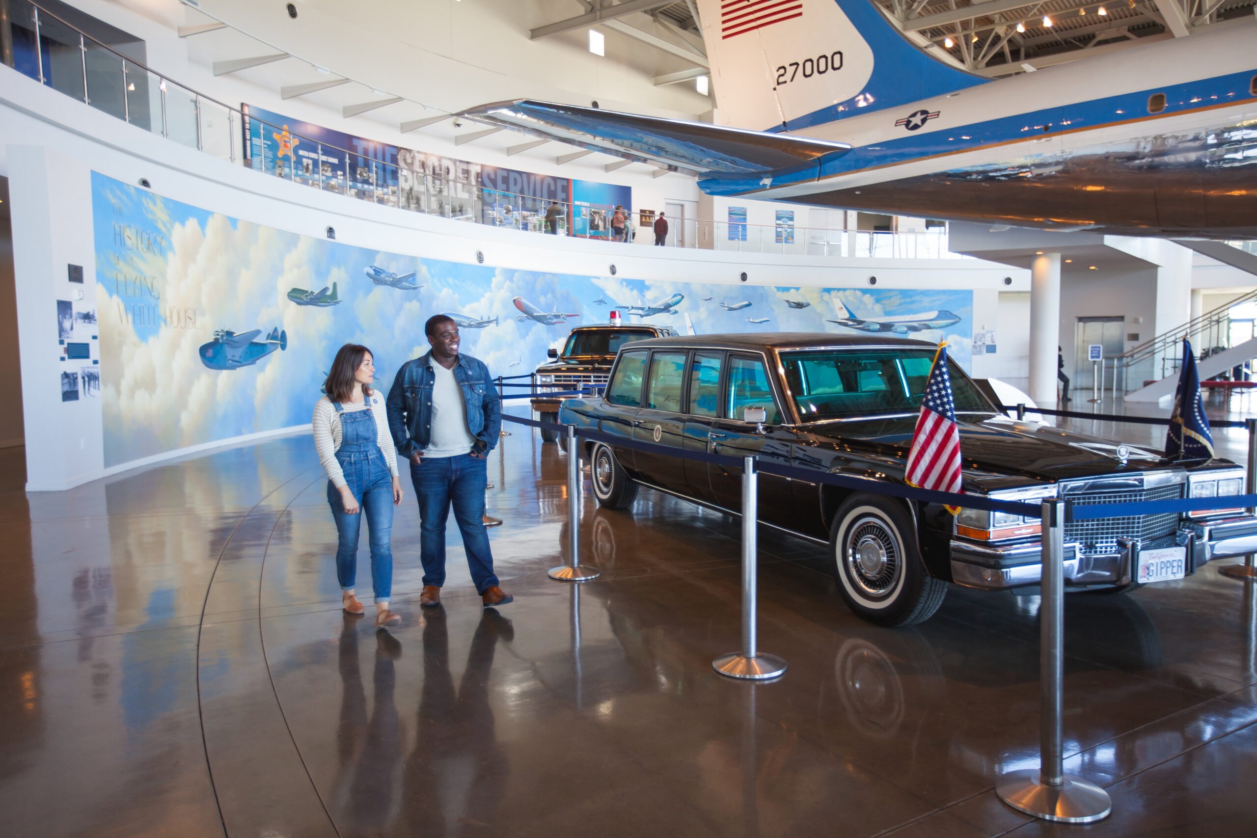 A couple walks in a modern museum with a presidential limousine displayed under an airplane wing.