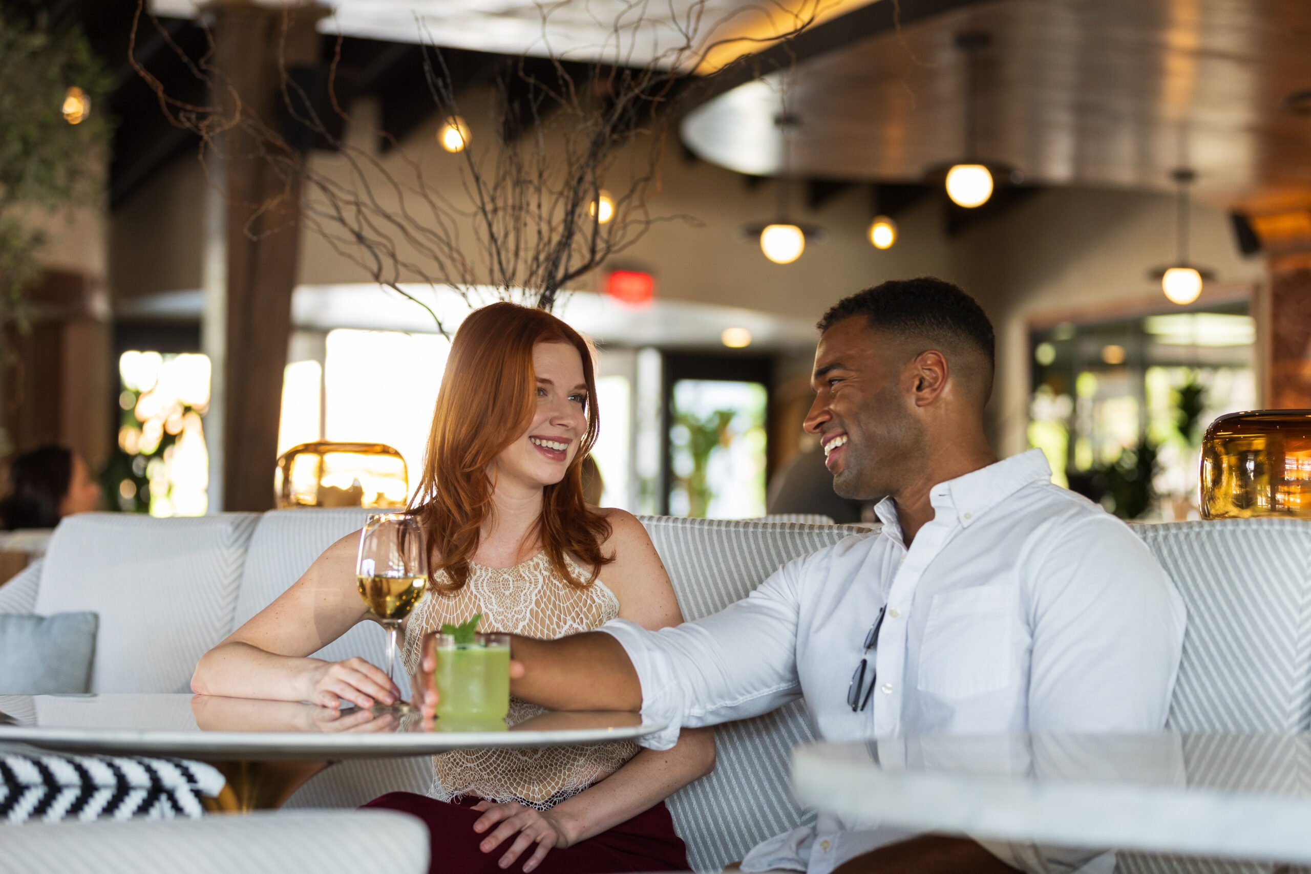 A man and a woman sit at a dining table, smiling and talking, each holding drinks.