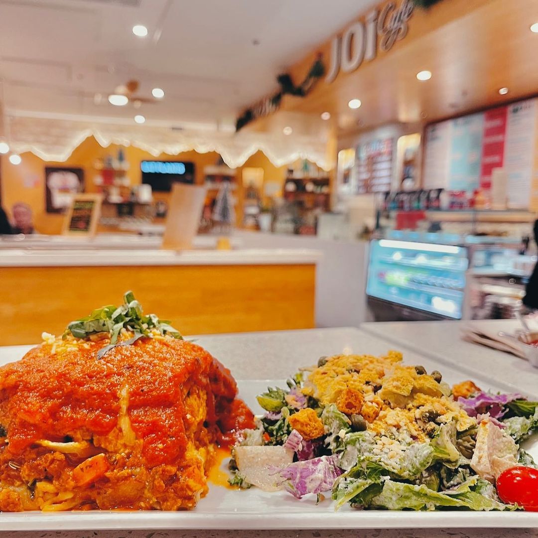 A plate of lasagna topped with tomato sauce and herbs sits beside a colorful salad with croutons in a bright, warmly lit café, featuring wood paneling and visible menu boards in Conejo Valley.
