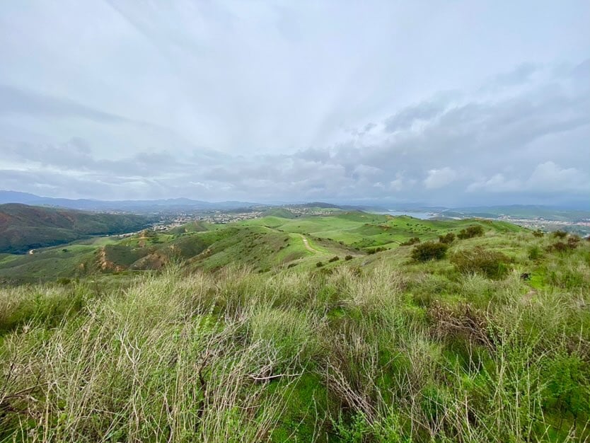 Rolling green hills stretch across the landscape under a cloudy sky, with distant mountains and a town visible on the horizon with a serene natural scene in Conejo Valley.