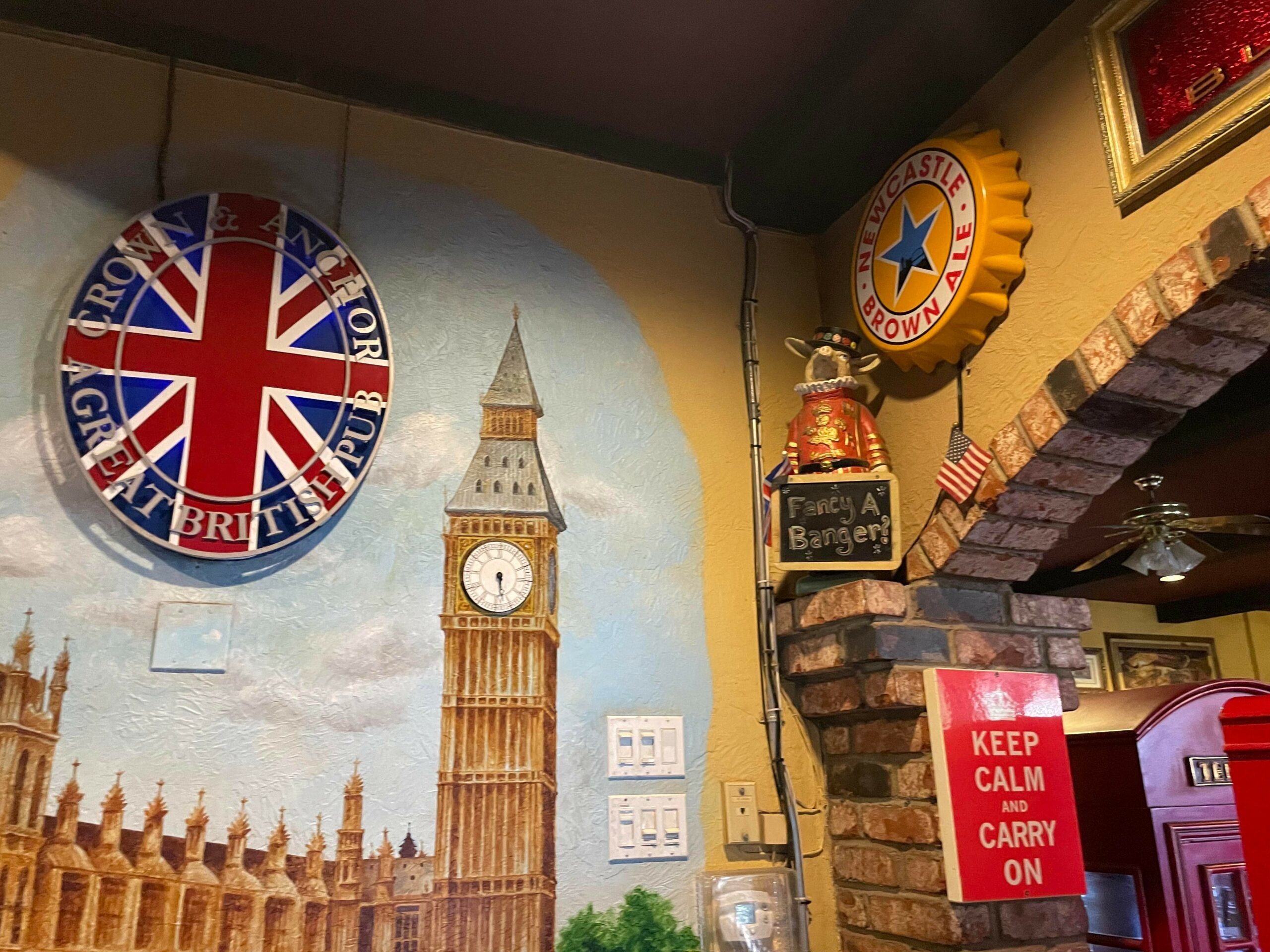 British pub decor featuring the Union Jack with "Crown & Anchor A Great British Pub," a mural of Big Ben, a Newcastle Brown Ale sign, a "Keep Calm and Carry On" poster, and a sign saying "Fancy a Banger?" in a cozy, themed setting in Conejo Valley.