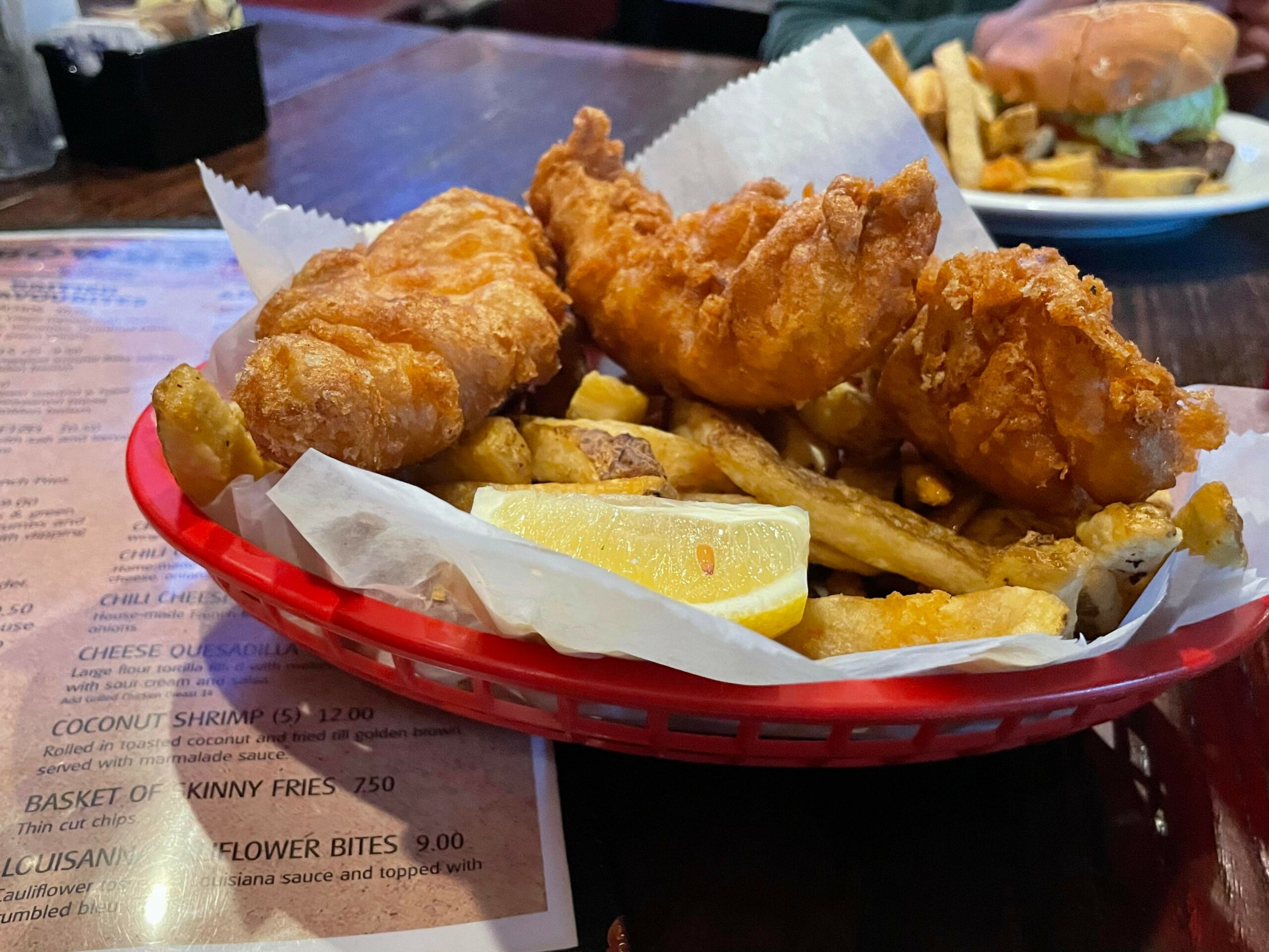 Basket of crispy fried fish and golden fries is served with lemon wedges on parchment paper; placed on a wooden table with a menu partially visible underneath in Conejo Valley.