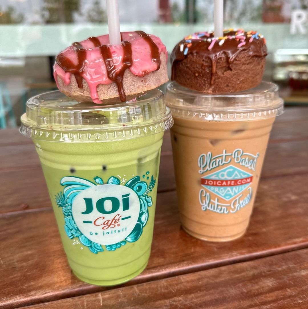 Two colorful iced drinks from Joi Café, one green and the other brown, topped with pink and chocolate-frosted donuts, rest on a wooden table. Text includes: “Joi Café” and “Plant-Based, Gluten-Free, joicafe.com in Conejo Valley.