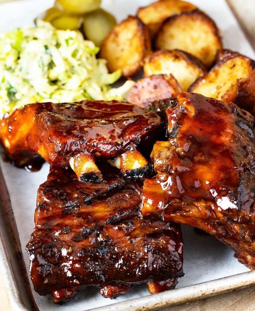 Barbecue ribs glisten with rich sauce on a tray, accompanied by crispy roasted potatoes and a serving of creamy coleslaw with a mouth-watering barbecue feast in Conejo Valley.
