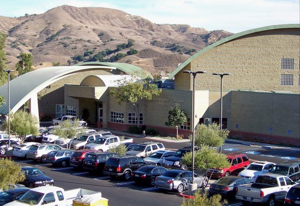 Cars parked in front of a modern building with arched roof labeled "Calabasas Community Center." Hilly landscape in the background, suggesting a suburban environment in Conejo Valley.