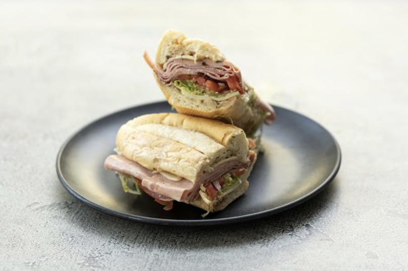 Sub sandwich sliced in half, revealing layers of deli meats, lettuce, and tomato, sits on a black plate.