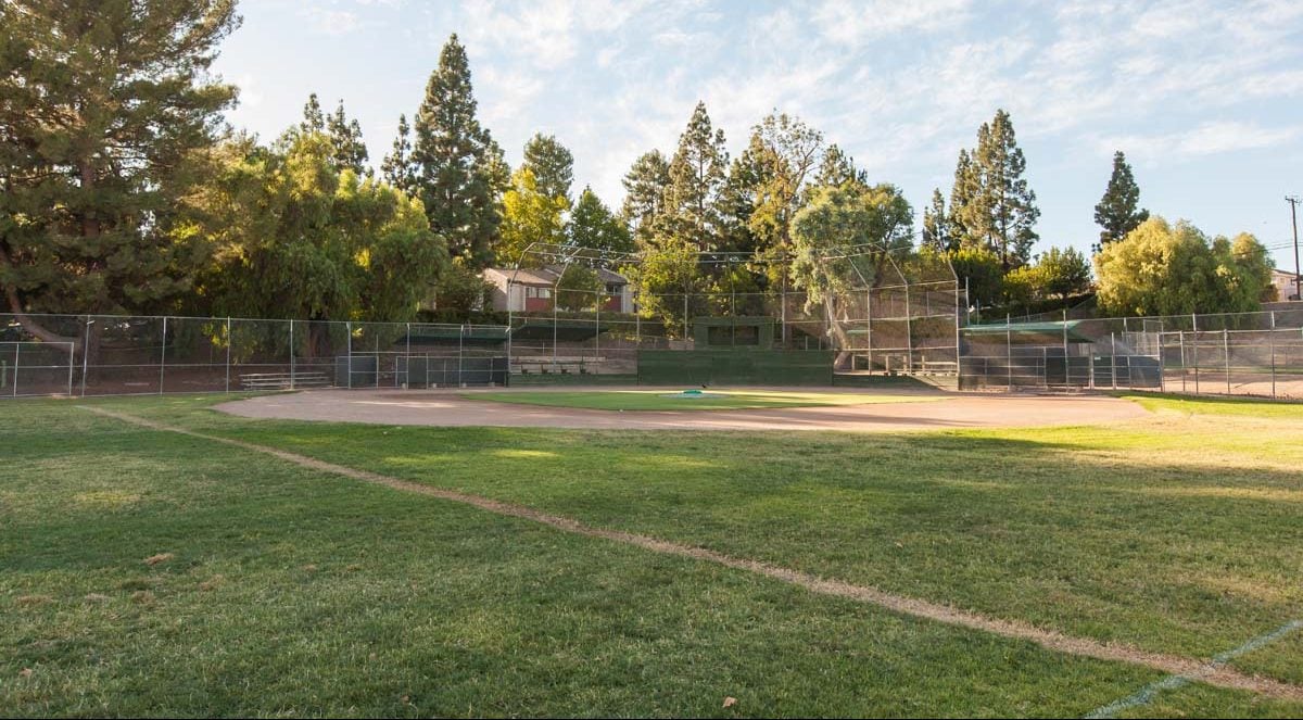 Baseball field sits empty, surrounded by lush green grass and tall trees, with a clear sky overhead and residential houses visible in the background, providing a serene park setting in Conejo Valley.