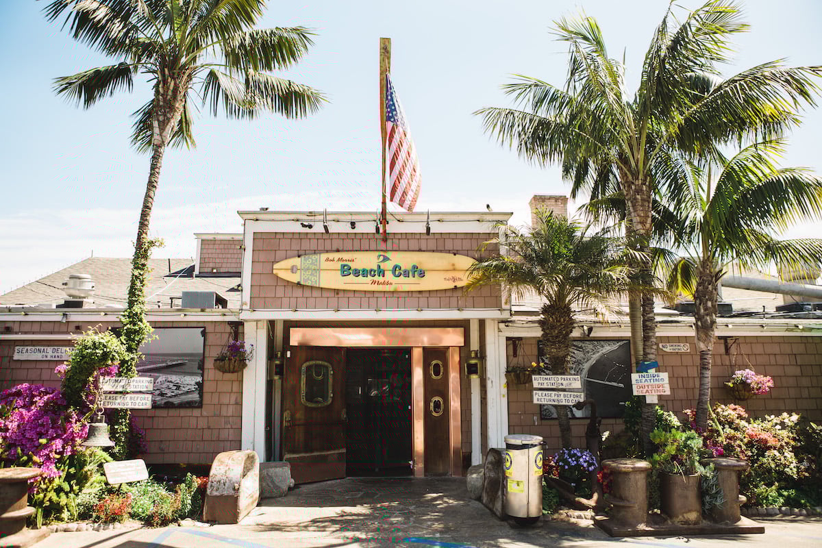 Beach café entrance beneath an American flag, surrounded by palm trees and vibrant flowers.