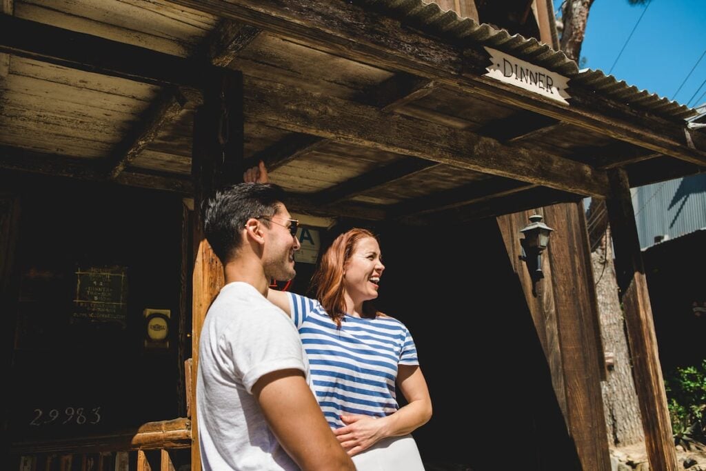 A smiling couple stands under an old wooden structure with a "DINNER" sign, suggesting an outdoor dining scene.