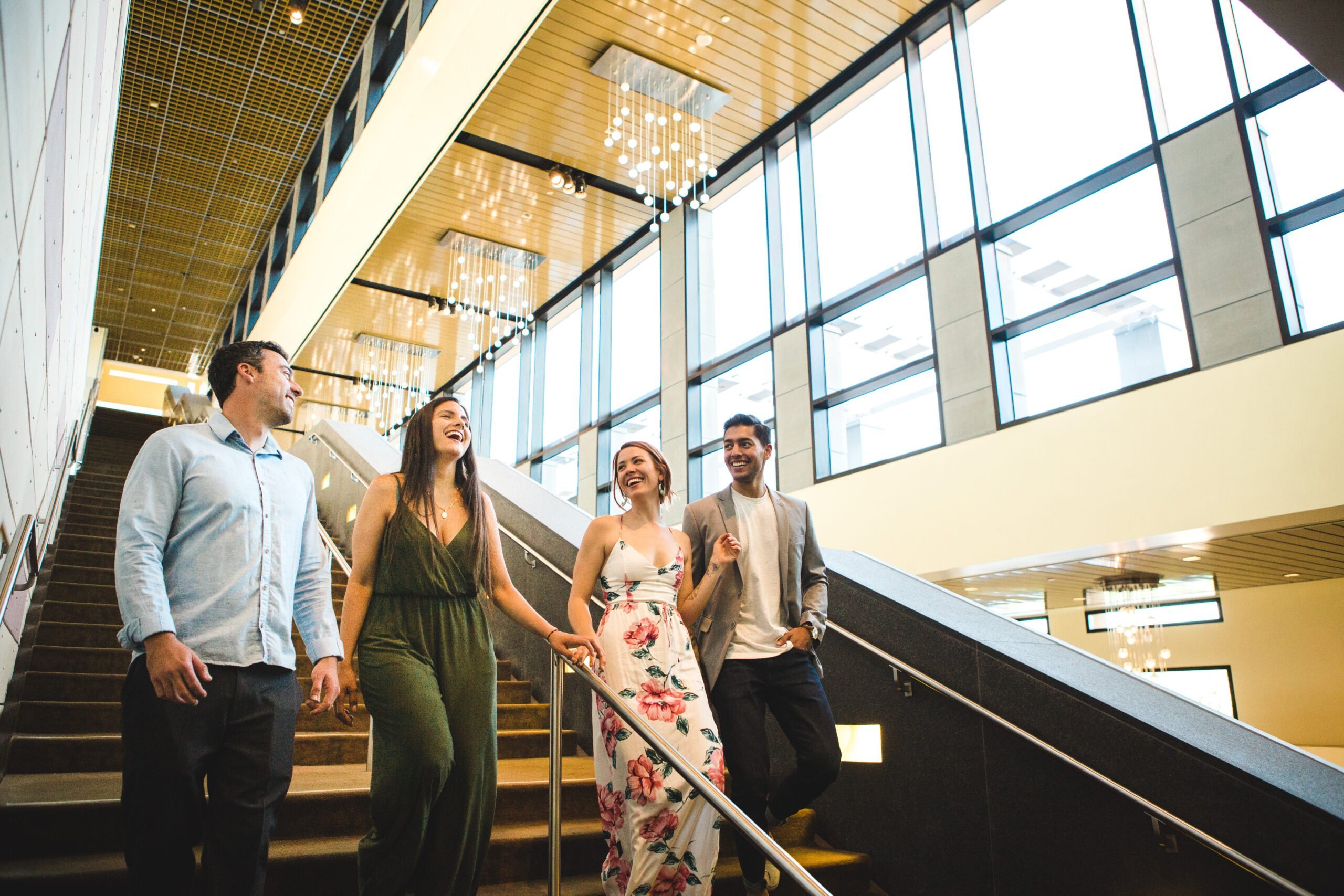 Group of four people walking down a modern staircase, laughing and chatting, under sleek lighting fixtures, with large windows showcasing a bright, spacious interior in Conejo Valley.