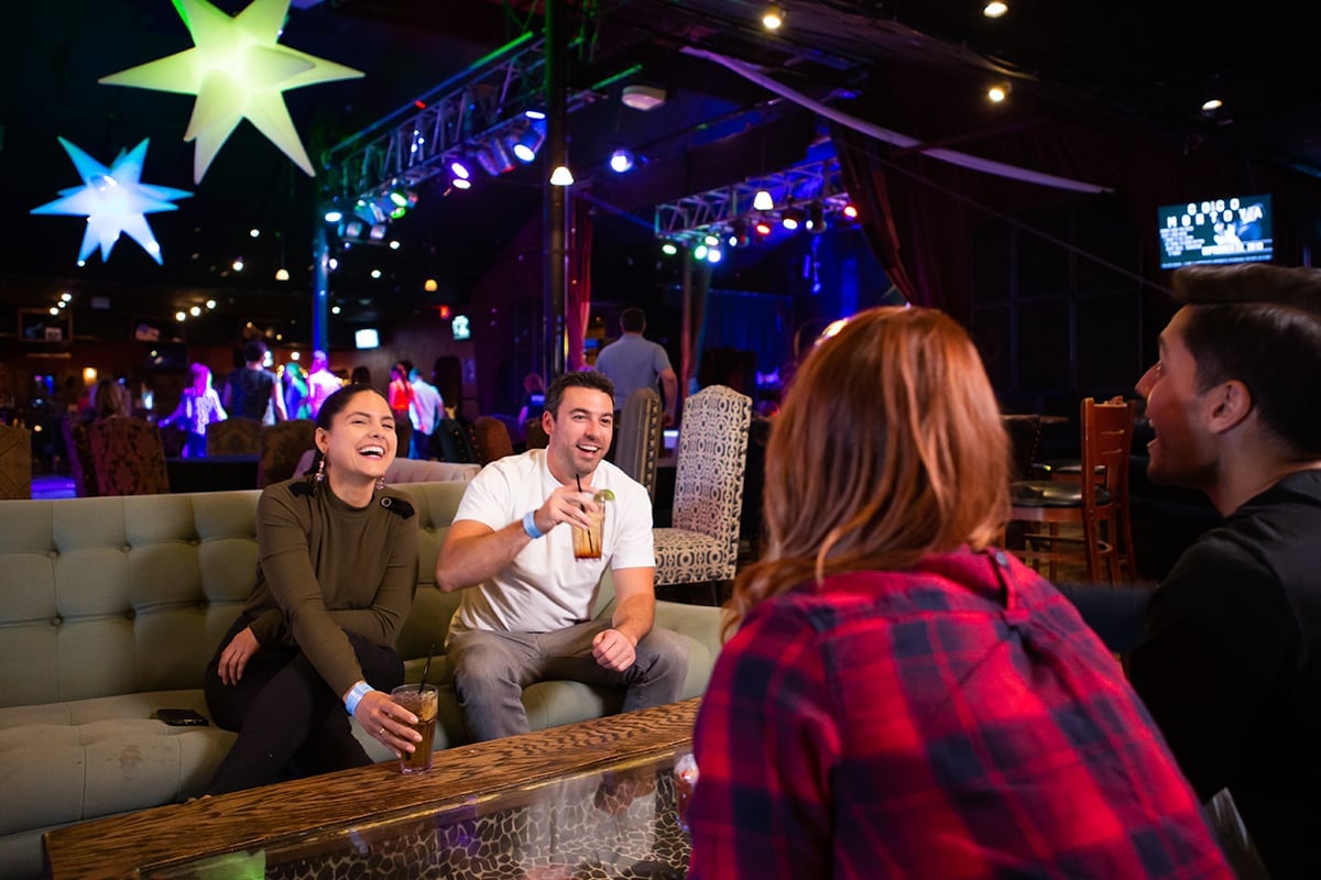 People laughing and drinking on a couch in a vibrant, colorful nightclub with star-shaped lights overhead; a dance floor and stage visible in the background in Conejo Valley.