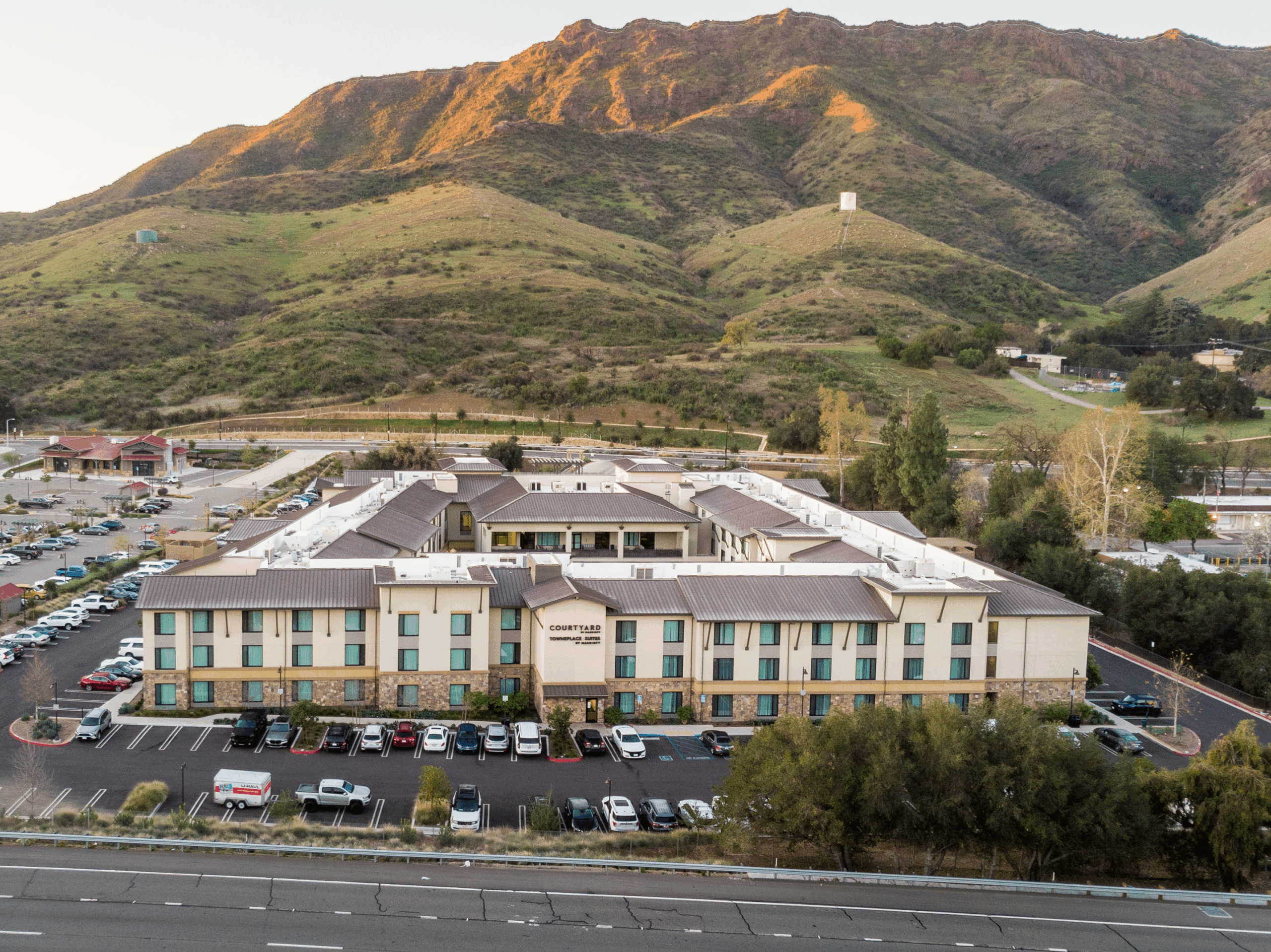 Courtyard hotel, surrounded by parked cars, is set against a backdrop of green, rolling hills.