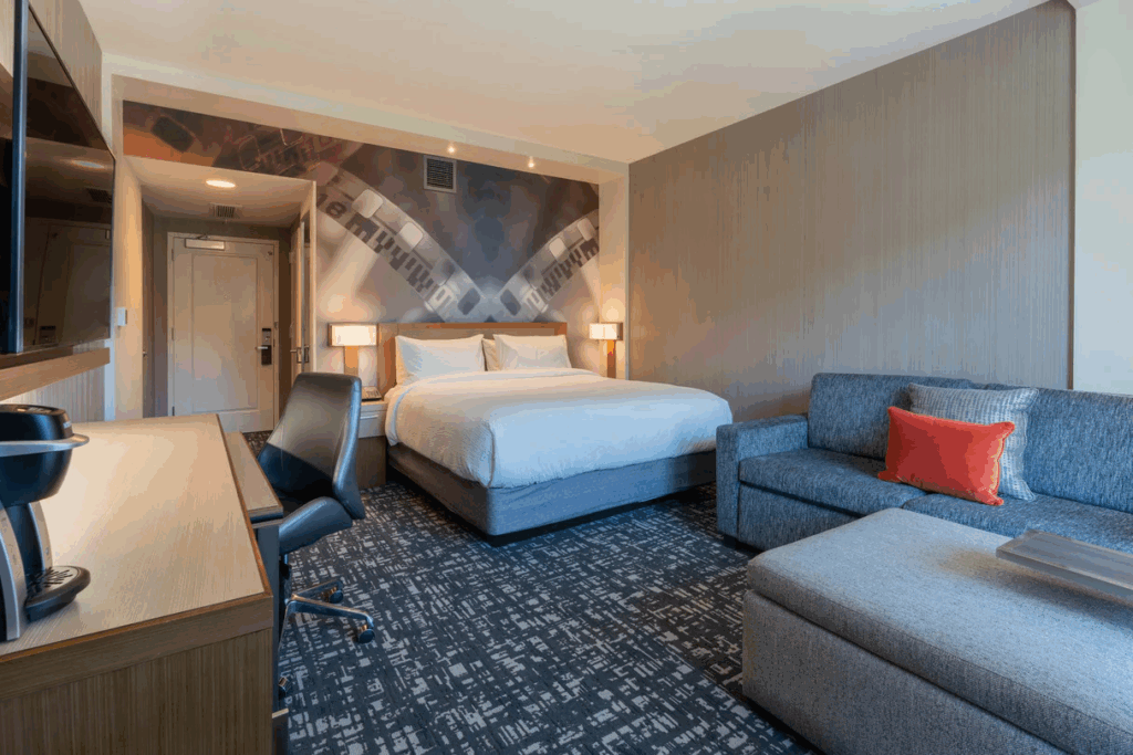 Modern hotel room featuring a comfortable double bed with white linens, a stylish gray sofa with a red pillow, and a work desk with a coffee maker. The artful wall displays abstract film imagery in Conejo Valley.