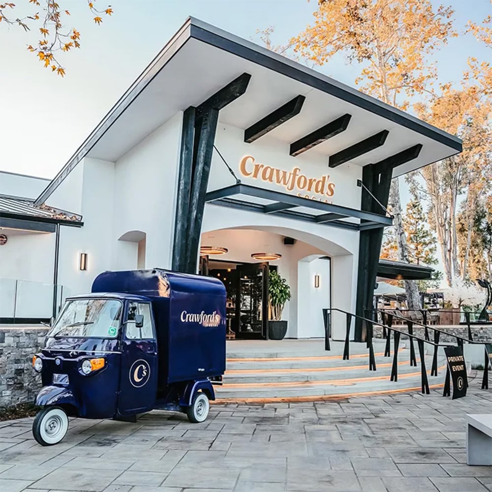 Blue three-wheeled vehicle parked in front of a modern building entrance. "Crawford's" is inscribed above the entrance and on the vehicle.