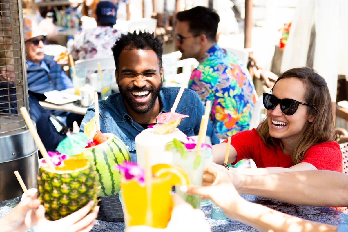 Friends enjoy colorful tropical drinks in hollowed fruits, laughing and clinking glasses outdoors, surrounded by a festive atmosphere and vibrant attire, perfect for a summer celebration in Conejo Valley.