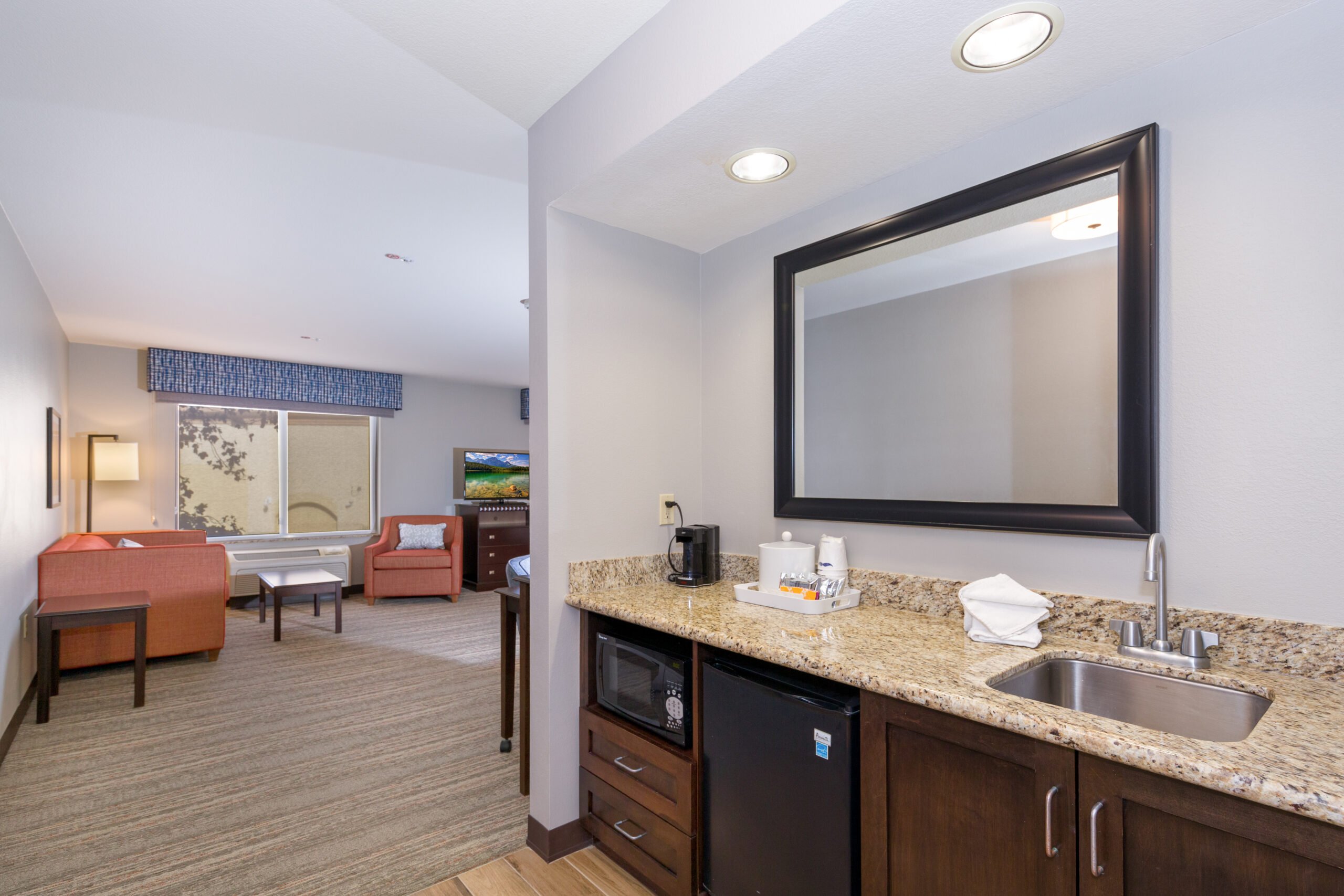 Kitchenette area with granite countertop, microwave, and mini-fridge, next to a cozy living space featuring orange seating, TV, and large window under a blue valance in a hotel suite in Conejo Valley.