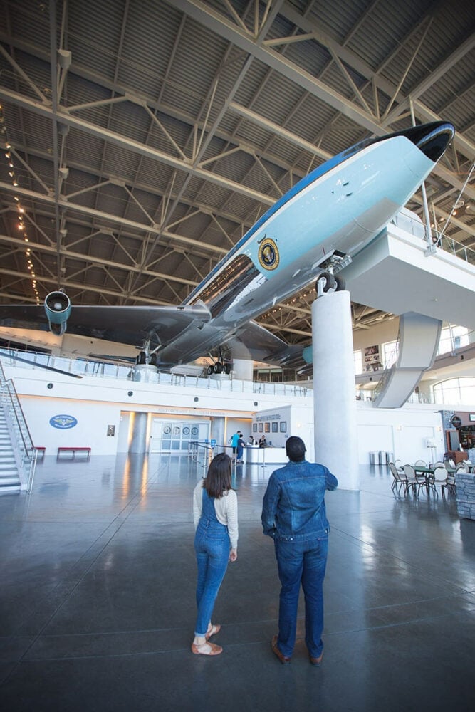 Large airplane suspended in a museum, viewed by two people standing on a glossy floor. The aircraft features the presidential seal.