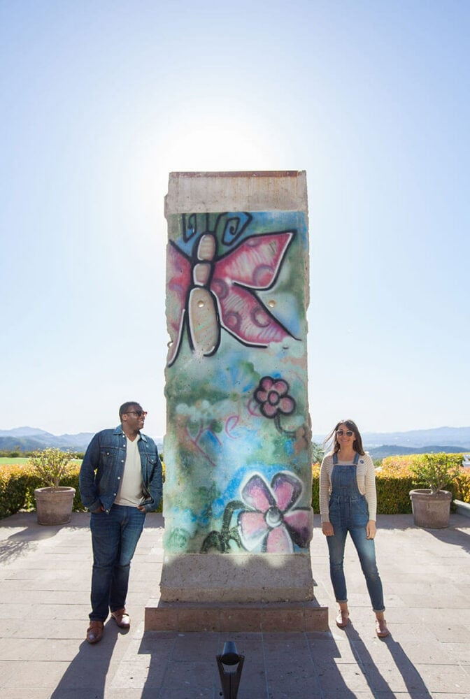 Concrete pillar with colorful butterfly and flower graffiti stands between two people on a sunny patio, surrounded by potted plants and distant mountains in Conejo Valley.