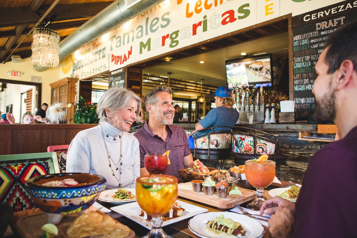 Group of people enjoying a meal at a vibrant Mexican restaurant. Drinks and colorful dishes are on the table.