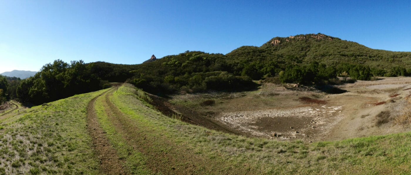 Grass-covered trail curves alongside a dry creek bed, surrounded by dense green shrubbery and rolling hills under a clear blue sky.