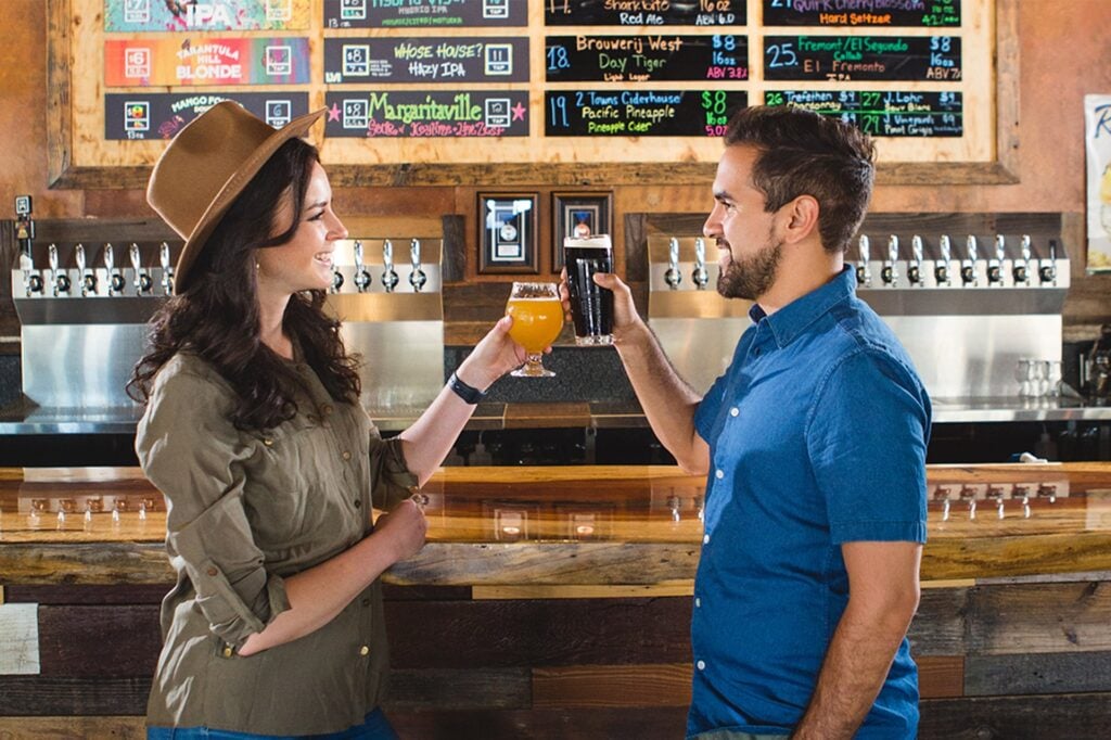 Two people clink beer glasses in a lively bar setting. A variety of beer taps are mounted on a wooden wall.