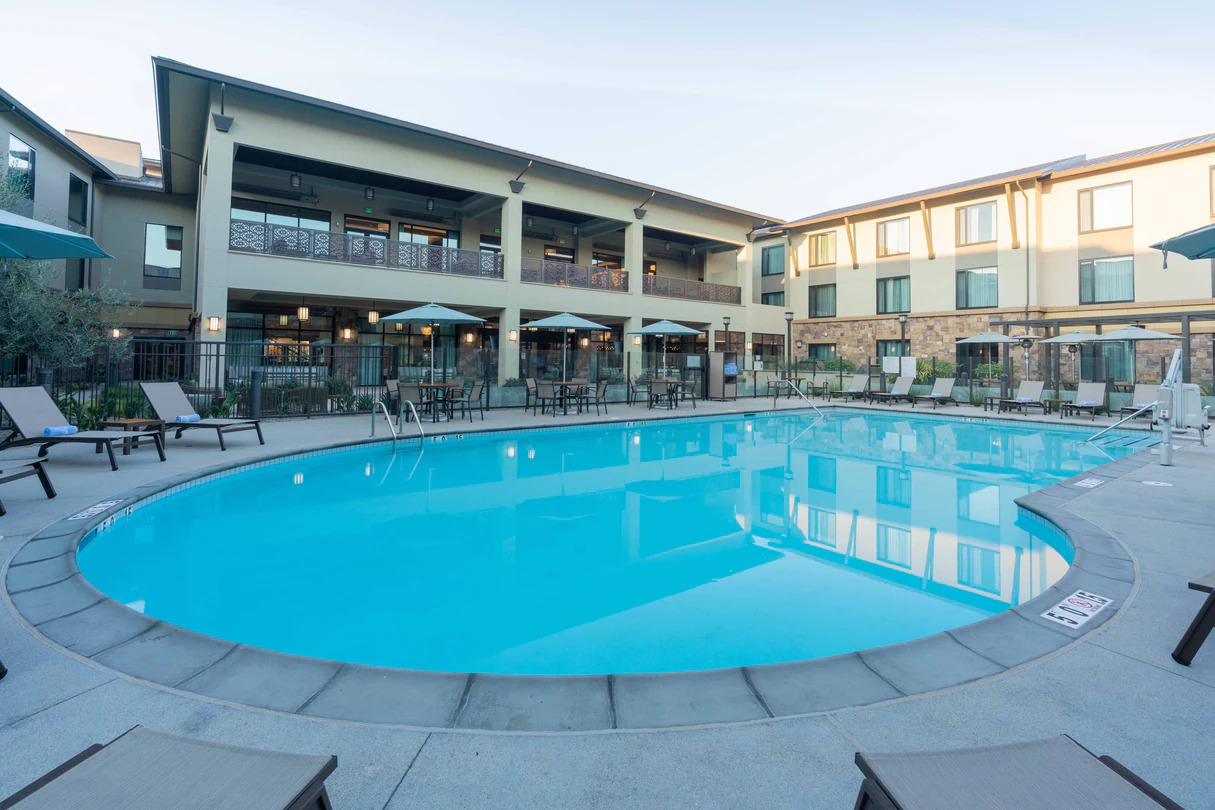 Outdoor swimming pool with clear blue water, surrounded by lounge chairs and umbrellas, located within a hotel courtyard.