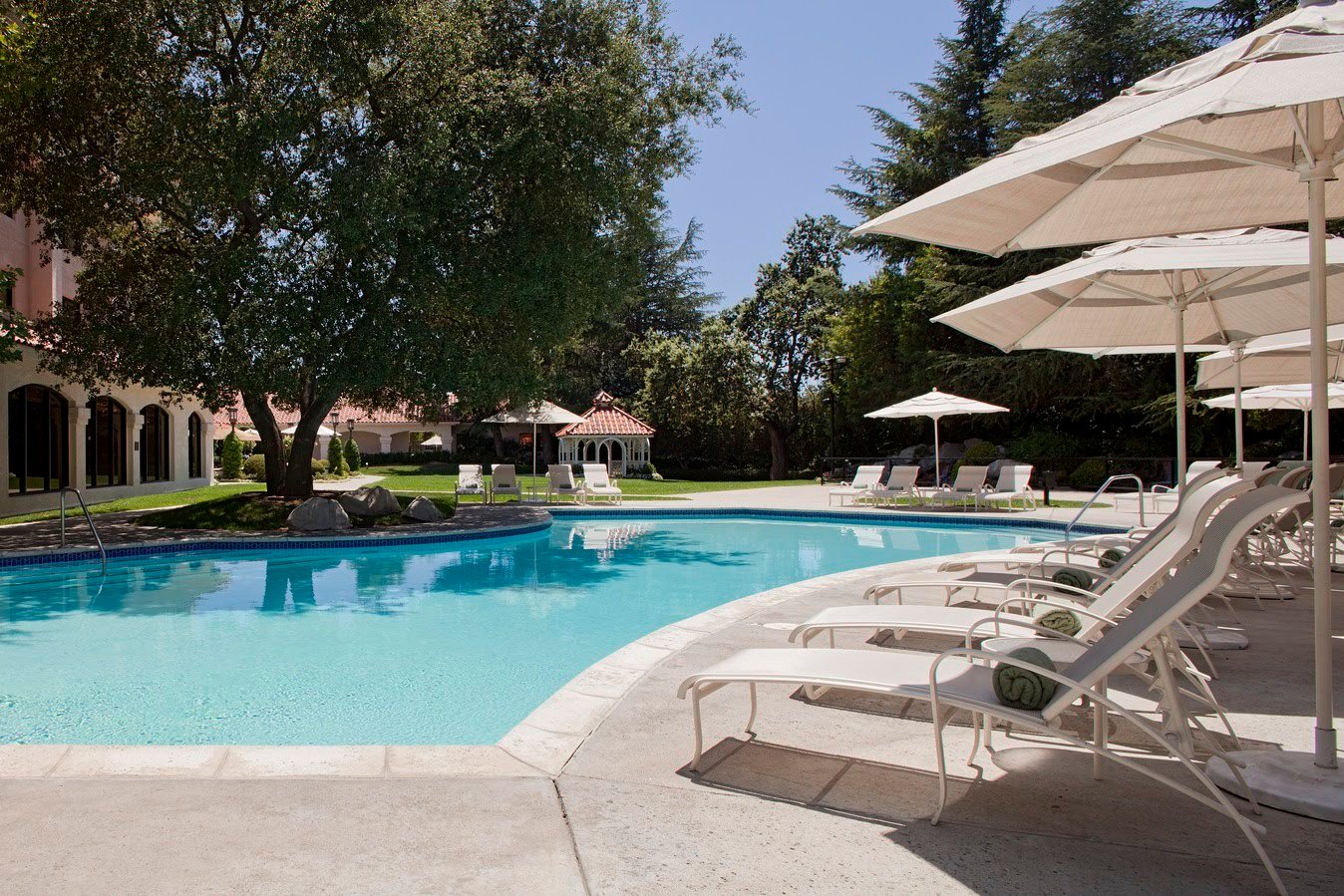Lounge chairs with towels sit under umbrellas by a clear swimming pool. A large tree provides shade, surrounded by greenery and a gazebo.