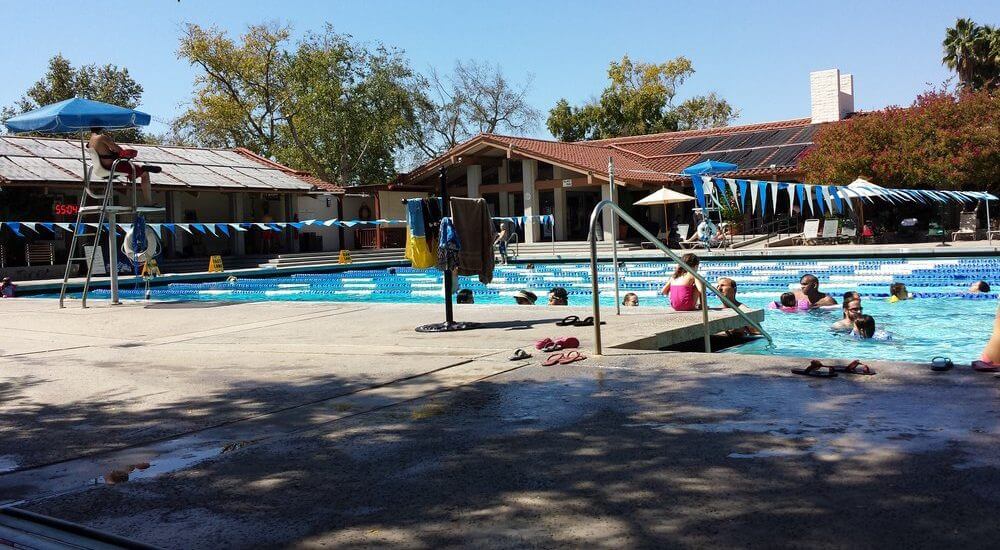 Public swimming pool with several people enjoying the water. A lifeguard sits under an umbrella.